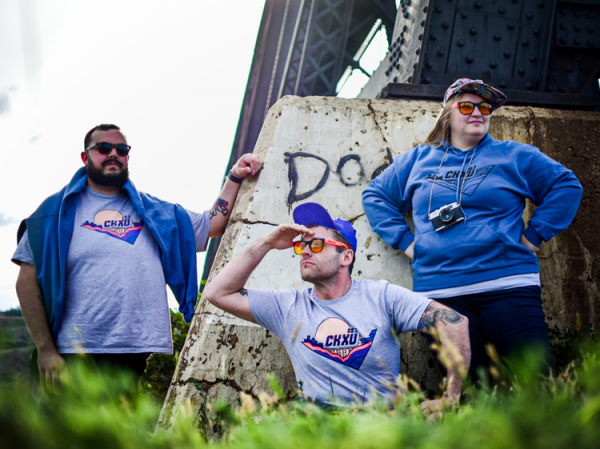 Pictured are three CKXU Staff members, posed as if they were tourists with hats, yellow-lens sunglasses, and a camera. Two of them are wearing the grey FUNdrive 2022 t-shirts with colored logo, the other is wearing the blue FUNdrive 2022 Hoodie with black linework logo. The background includes parts of the historic Lethbridge High Level Bridge.
